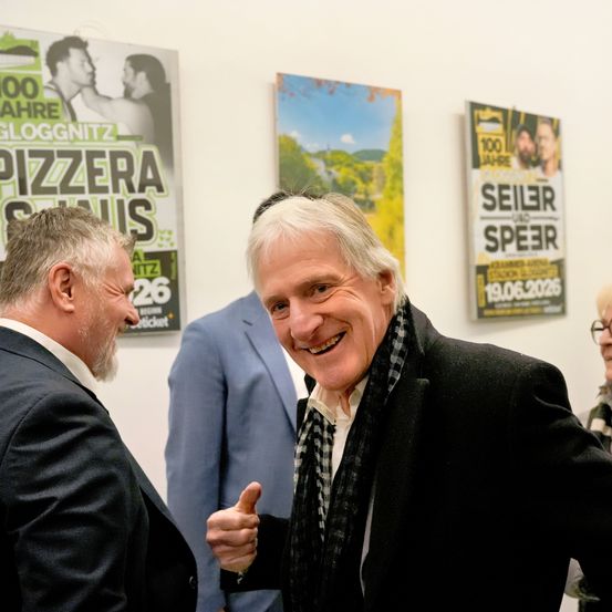 Zwei Männer in Anzügen unterhalten sich in der Nähe einer Wand mit Plakaten. Ein Mann gibt einen Daumen hoch. Eine weitere Person in einem blauen Anzug steht hinter ihnen.