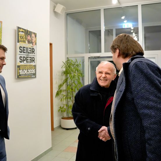Drei Männer unterhalten sich in einem Flur mit einem Plakat an der Wand, einer Pflanze in einem weißen Topf und Glastüren.
