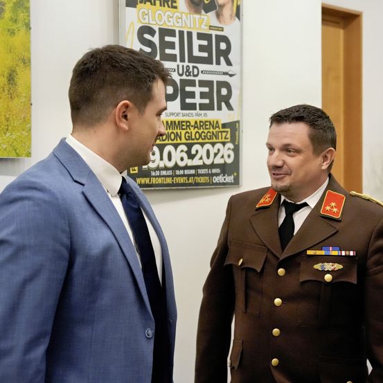 Zwei Männer unterhalten sich; einer in Anzug und Krawatte, der andere in Uniform. Ein Plakat hinter ihnen bewirbt ein Konzert am 9. Juni 2026.