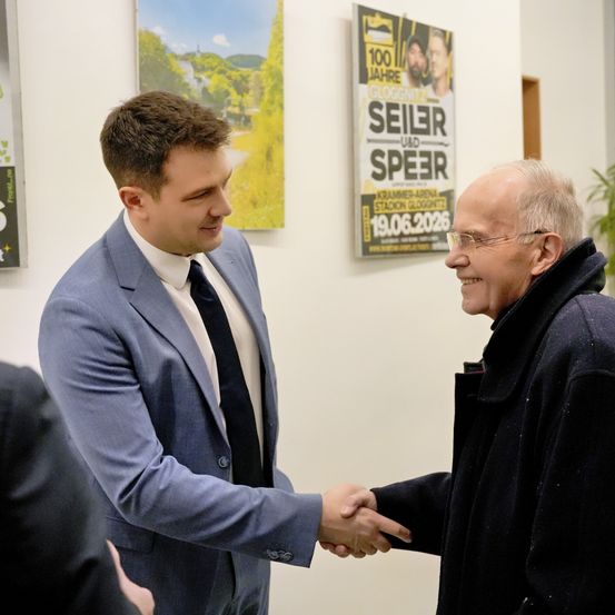 Zwei Männer schütteln sich die Hände in einem Raum. Der Mann im Anzug trägt eine Brille und lächelt. Der andere Mann ist älter, trägt einen Mantel und hat einen Bart. Hinter ihnen befindet sich ein Plakat mit den Worten 'Seiler und Speer'.