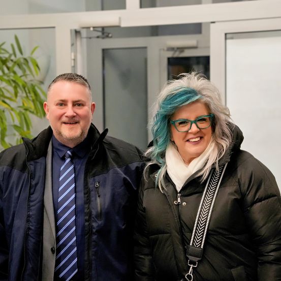 Ein Mann und eine Frau stehen eng beieinander und lächeln für ein Foto. Der Mann trägt ein blaues Hemd mit einem gestreiften Krawatte und einer Jacke. Die Frau hat blaue Haare, eine Brille und einen weißen Schal. Sie stehen vor einem Gebäude mit Glastüren.