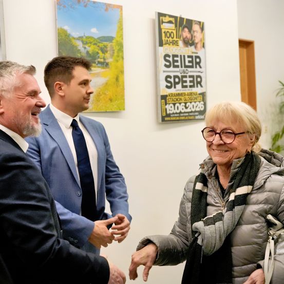 Drei Männer und eine Frau stehen vor einer Wand. Zwei Männer schütteln Hände, während die anderen beiden lächeln. Dahinter ist ein Plakat für eine 'Seiler und Speer'-Veranstaltung.