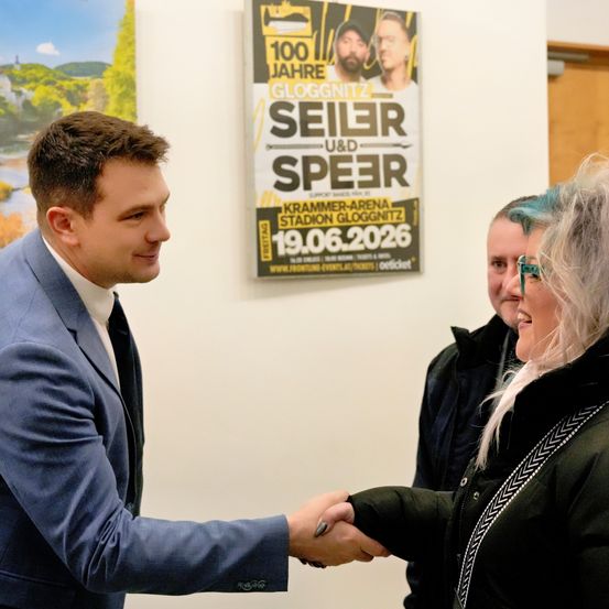 Ein Mann in einem blauen Anzug schüttelt die Hand einer Frau in einer schwarzen Jacke. Ein Mann mit blauen Haaren steht hinter ihnen. Sie sind vor einem Plakat für ein Konzert.