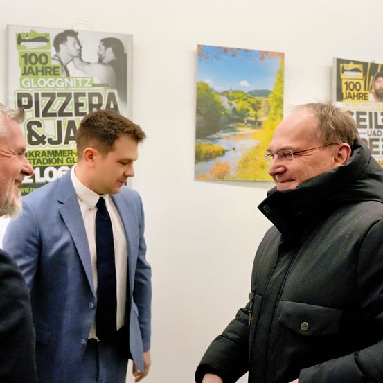 Drei Männer unterhalten sich in einem Raum mit Postern an der Wand. Ein Poster trägt die Aufschrift '100 Jahre Gloggnitzer'. Der Mann in der Mitte trägt Anzug und Krawatte.