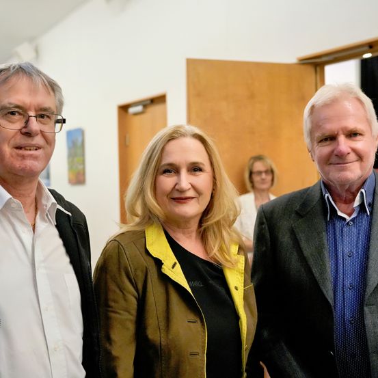 Drei Personen stehen zusammen in einem Raum. Der Mann auf der linken Seite trägt eine Brille und eine schwarze Jacke. Die Frau in der Mitte hat blonde Haare und trägt eine gelbe Jacke. Der Mann auf der rechten Seite trägt ein blaues Hemd und eine schwarze Jacke.