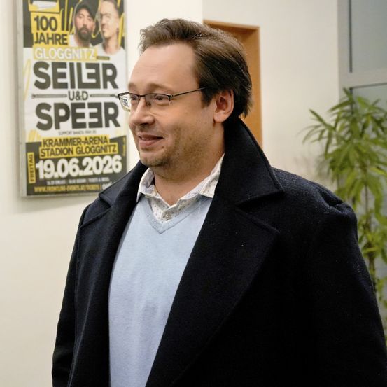 Ein Mann mit Brille und Bart steht lächelnd vor einem Plakat der Band Seiler und Speer.