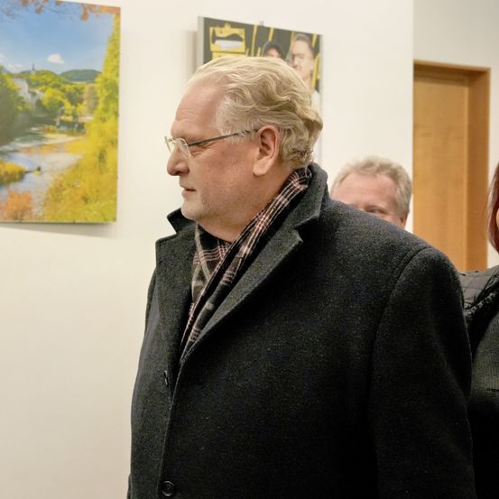 Ein Mann mit Brille und Mantel steht vor einer Wand mit Kunst und betrachtet ein Landschaftsgemälde. Ein anderer Mann hinter ihm schaut auf das Kunstwerk.
