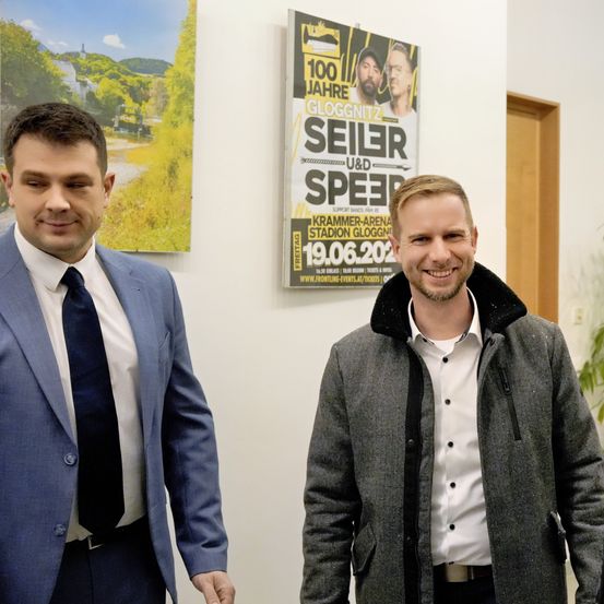 Zwei Männer stehen lächelnd in einem Büro, einer im Anzug und Krawatte, der andere im grauen Mantel. Ein Plakat hinter ihnen lautet '100 Jahre GLOGGNITZ SEILER SPEED' mit Datum und Veranstaltungsort.