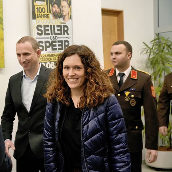 Eine lächelnde Frau in einer blauen Jacke steht zwischen zwei Männern, einer in Anzug und der andere in Uniform. Sie sind drinnen, mit Postern an der Wand und einem Topfplant im Hintergrund.