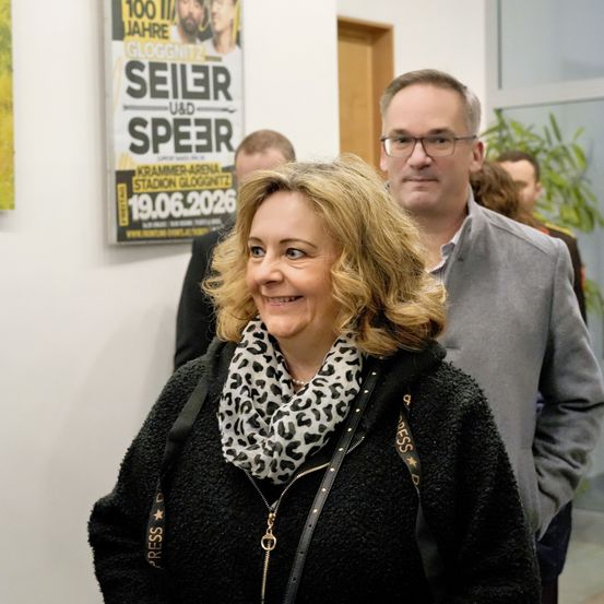 Eine Frau mit einem Leopardenmuster-Schal lächelt in die Kamera, neben einem Mann in einem grauen Hemd in einem Korridor mit einem Plakat, das ein Ereignis am 19. Juni 2026 ankündigt.