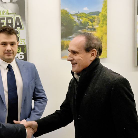 Zwei Männer in Anzügen schütteln sich die Hände in einer geschlossenen Umgebung. Hinter ihnen ist ein landschaftliches Poster.