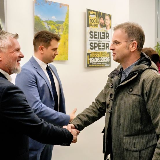 Drei Männer schütteln sich in einem Raum die Hände. Zwei tragen Anzüge, einer eine Jacke. Ein Poster mit den Worten 'Seiler und Speer' ist an der Wand.