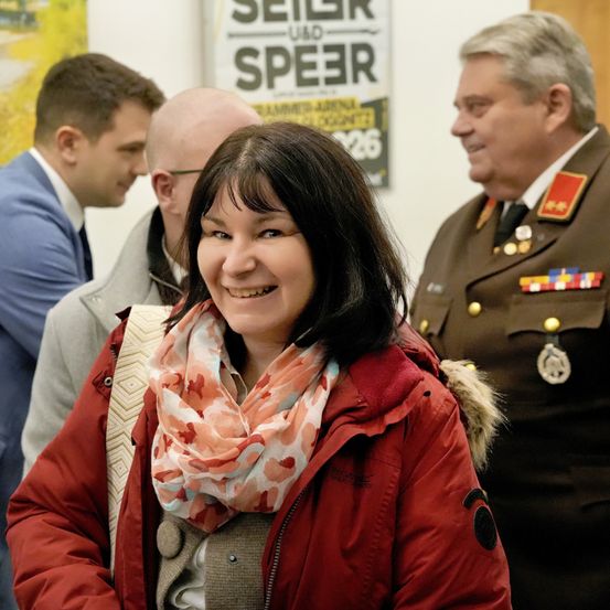 Eine Frau lächelt, trägt eine rote Jacke und steht zwischen zwei Männern in Anzügen und einem in Uniform. Sie stehen neben einem Plakat mit dem Wort Speer.