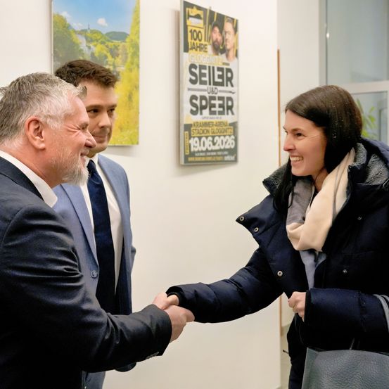 Drei Menschen schütteln sich die Hände in einer Innenumgebung. Ein Plakat auf Deutsch ist an der Wand sichtbar.