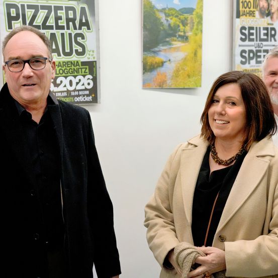 Drei Personen stehen vor einer weißen Wand mit Plakaten. Ein Mann mit Brille lächelt für die Kamera. Neben ihm steht eine Frau mit einer Halskette. Hinter ihnen ist ein Mann mit Bart.