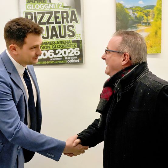 Zwei Männer schütteln sich die Hände in einem Raum mit einem Plakat, das ein Event in der Pizzara Jaus in Gloggnitz ankündigt.