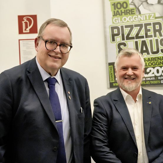 Zwei Männer in Anzügen lächeln für ein Foto in einem Büro. Hinter ihnen bewirbt ein Plakat '100 Jahre Gloggnitzer & Haus' mit einem Veranstaltungsdatum von 2020.