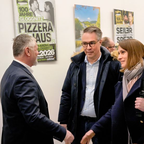 Drei Personen unterhalten sich in einem Raum mit Postern an der Wand, darunter eines für das Jahr 2026.