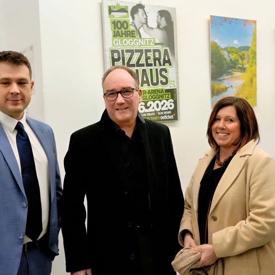 Drei Personen stehen vor einer Wand mit zwei Plakaten. Ein Mann in einem schwarzen Anzug und mit Brille steht zwischen einem lächelnden Mann und einer Frau. Die Plakate sind für eine 'Pizzera Haus' Veranstaltung.