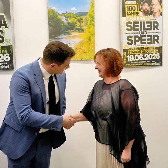 Ein Mann und eine Frau schütteln sich die Hände vor einer Wand mit Plakaten. Der Mann trägt einen blauen Anzug, und die Frau trägt ein schwarzes Kleid.