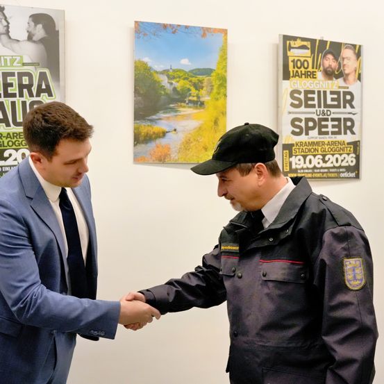 Ein Mann in einem blauen Anzug und Krawatte schüttelt die Hand eines Sicherheitsbeamten in einer schwarzen Jacke mit rotem Streifen und Abzeichen. Hinter ihnen befinden sich Poster eines Flusses und Berge sowie ein Veranstaltungsposter für ein Konzert.