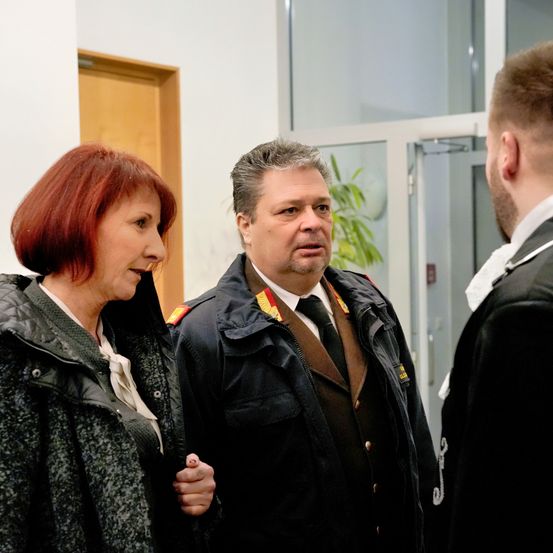 Eine Frau mit kurzen roten Haaren und ein Mann in Uniform unterhalten sich mit einer dritten Person in formeller Kleidung.