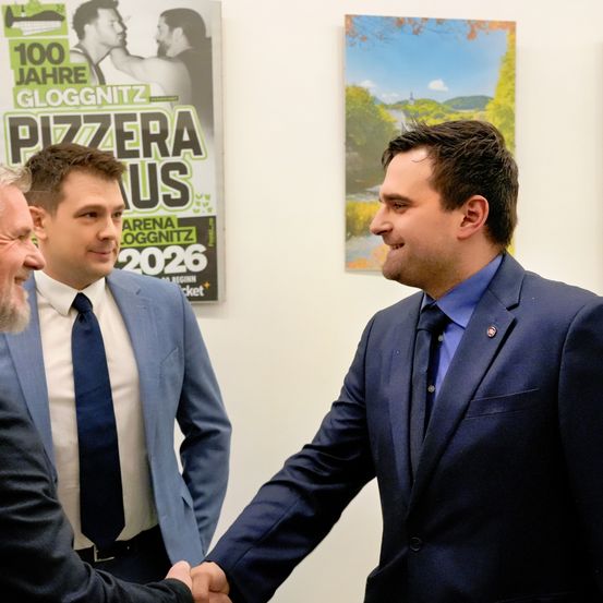 Drei Männer in Anzügen schütteln sich die Hände in einem Büro, mit einem Plakat für '100 Jahre Gloggnitz' und einem Landschaftsbild im Hintergrund.