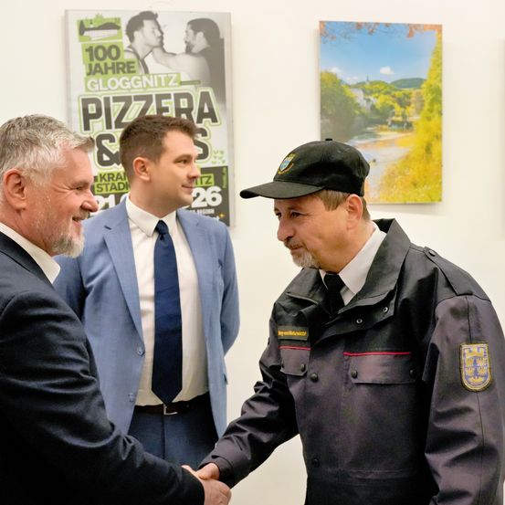 Drei Männer in formeller Kleidung schütteln sich die Hände. Ein Mann trägt eine schwarze Uniform und einen Hut. Sie stehen vor einer Wand mit Postern.