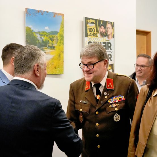 Eine Gruppe von Menschen steht in einem Innenraum. Ein Mann in Uniform lächelt und scheint mit dem Mann im Anzug zu sprechen. Hinter ihnen hängen zwei Poster an der Wand.