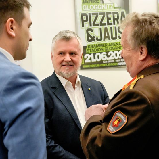 Drei Männer stehen in einem Raum. Einer ist in Uniform, ein anderer in einem Anzug. Hinter ihnen ist ein Schild mit Pizzara & Jaus und einem Datum.