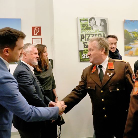 Ein Soldat in Uniform schüttelt einem anderen Mann die Hand. Hinter ihnen stehen drei Personen in der Nähe einer Wand mit zwei Plakaten.