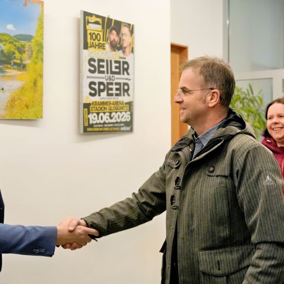 Zwei Männer schütteln sich die Hände in einer geschlossenen Umgebung. Ein Mann trägt eine Brille und eine grüne Jacke. Hinter ihnen lächelt eine Frau. Ein Plakat, das 'Seiler und Speer' bewirbt, ist an der Wand sichtbar.