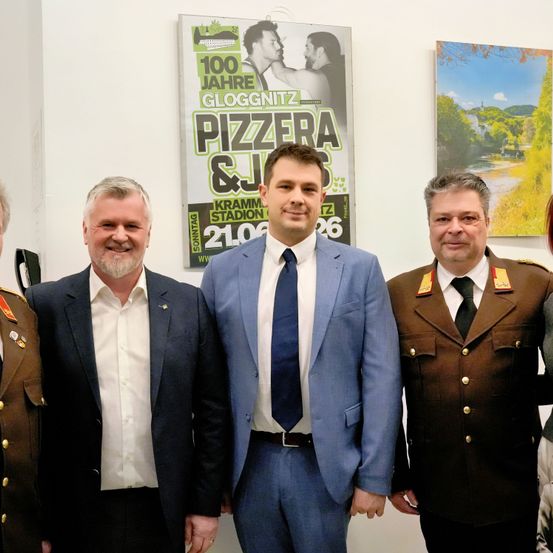 Vier Männer in Anzügen stehen vor einer weißen Wand mit zwei Postern, eines zeigt ein Paar und die Worte '100 Jahre Gloggnitzer Pizzeria & J.' Das andere Poster zeigt eine landschaftliche Ansicht mit einem Fluss und einer Brücke.