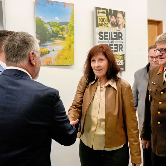 Eine Frau in einer braunen Lederjacke begrüßt einen Mann im Anzug in einem Raum mit Postern an der Wand.
