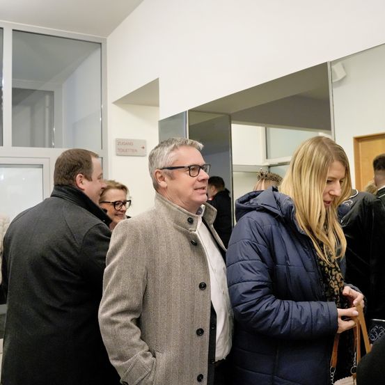 Im Inneren unterhalten sich mehrere Personen. Ein Mann mit Brille und Mantel steht neben einer Frau mit blonden Haaren in einer blauen Jacke. Ein Schild weist auf Toiletten hin.