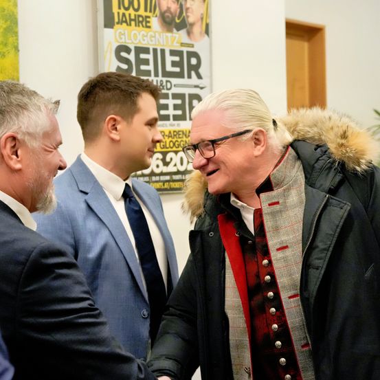 Drei Männer in Anzügen und mit Brillen, einer in einem Pelzmantel, unterhalten sich. Im Hintergrund befindet sich ein Plakat mit einem bärtigen Mann und den Worten 'GLOGGNITZ' und '100 Jahre'.