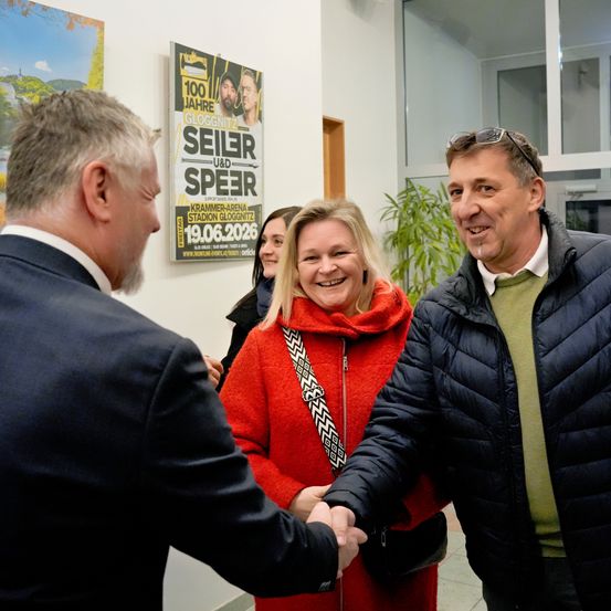 Drei Menschen stehen in einem Flur, lächeln und schütteln Hände. Eine Frau in einem roten Mantel steht in der Mitte, mit einem Mann rechts von ihr, der eine schwarze Jacke trägt. Hinter ihnen befindet sich ein Plakat für die Veranstaltung "100 Jahre Gloggnitzer".
