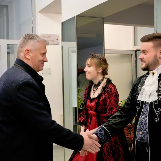 Zwei Männer und eine Frau in einem roten Kleid schütteln sich die Hände. Die Frau trägt eine Krone. Sie stehen in einem Raum, in der Nähe einer Glastür mit einem Schild, das 'Zugang' besagt.