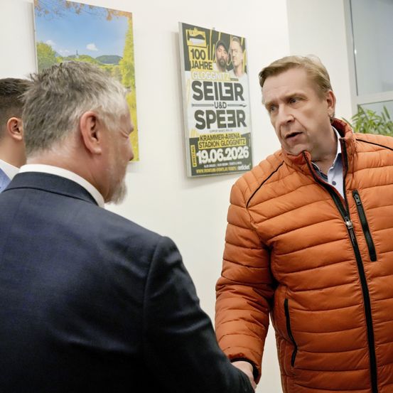 Zwei Männer schütteln sich die Hände in einem Büro. Einer trägt eine braune Jacke, der andere einen dunklen Anzug. Hinter ihnen ist ein Plakat mit einem Datum und Personen.