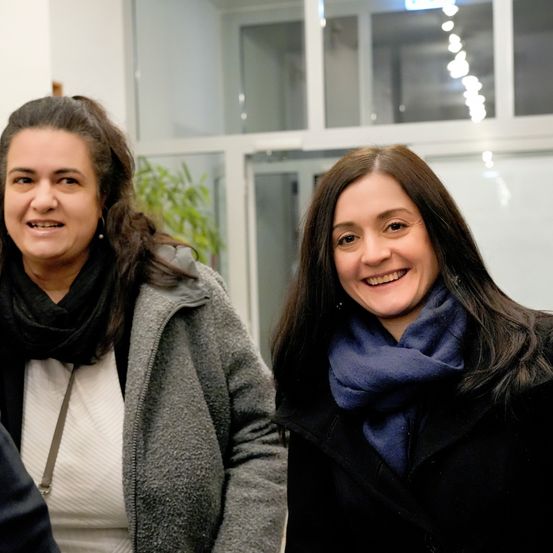 Zwei Frauen lächeln in einem Gebäude mit Glaswänden. Die Frau auf der linken Seite trägt eine graue Jacke und einen schwarzen Schal. Die Frau auf der rechten Seite trägt einen schwarzen Mantel und einen blauen Schal.