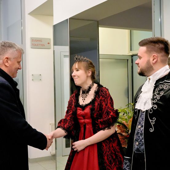 Ein Mann in einem schwarzen Mantel schüttelt die Hand einer Frau in einem roten Kleid und Krone, während ein Mann in einem schwarzen und weißen Kostüm zuschaut. Sie befinden sich in einem Flur mit einer weißen Wand und einem Schild für Toiletten.