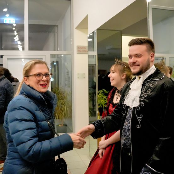 Eine Frau in einer blauen Jacke schüttelt einem Mann in einem Kostüm die Hand in einem Gebäude. Dahinter schaut eine Frau in einem roten Kleid und Krone zu. Ein Schild mit der Aufschrift 'Zugang Toiletten' ist sichtbar.