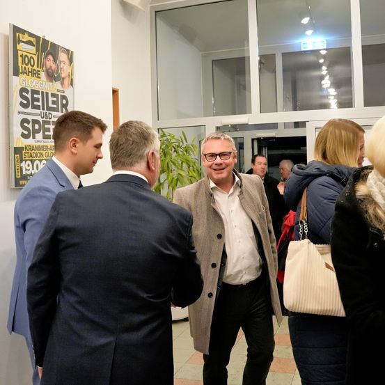 Mehrere Männer in Anzügen unterhalten sich in einem Flur, ein Mann mit Brille lächelt und blickt nach vorne. Ein Poster an der Wand trägt den Text 'Seiler' mit einem Preisschild.