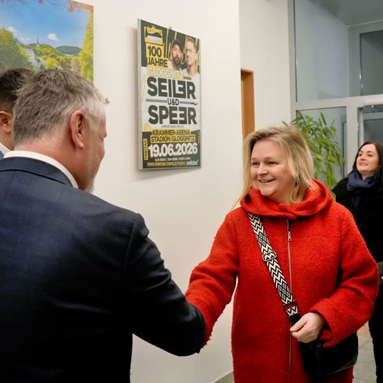 Eine Frau in einem roten Mantel lächelt und schüttelt einem Mann im Anzug die Hand. Hinter ihnen befindet sich ein Plakat für ein Konzert.