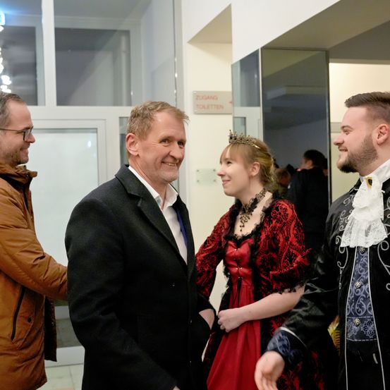 Vier Menschen sind drinnen, drei Männer und eine Frau. Die Frau trägt ein rotes Kleid und eine Krone, während die Männer Anzüge tragen. Sie scheinen zu unterhalten. Hinter ihnen befindet sich eine Tür mit einem Schild, das 'Zugang Toiletten' besagt.