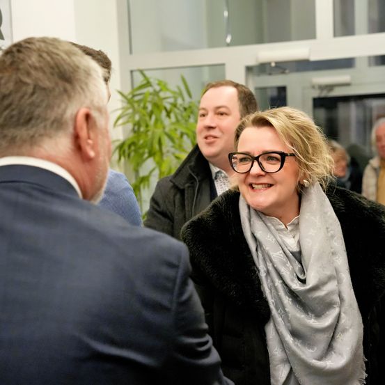 Eine Gruppe von Menschen steht in einem Raum, zwei Frauen unterhalten sich, während zwei Männer ein Gespräch führen. Eine Frau mit Brille und Schal lächelt.