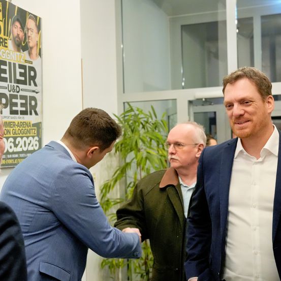 Drei Männer in Anzugkleidung stehen vor einer Glastür und unterhalten sich, wobei ein Plakat mit zwei Männern im Hintergrund zu sehen ist.