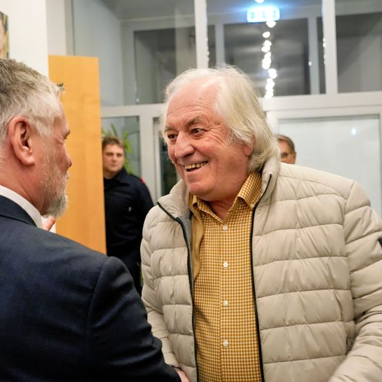 Zwei Männer lächeln und schütteln sich die Hände in einem Raum mit Glasfenstern und einer Holztür. Der Mann im gelben karierten Hemd trägt eine beigefarbene Daunenjacke.