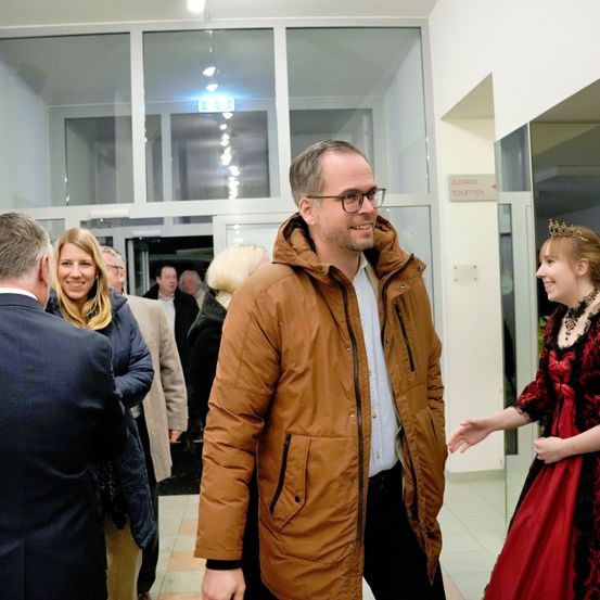 Ein Mann mit Brille und brauner Jacke unterhält sich mit einer Frau in einem roten Kleid in einem Flur mit Glaswänden.
