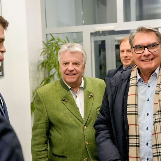 Drei Männer in einem Flur, einer trägt eine grüne Jacke und ein anderer trägt eine Brille und einen Schal.